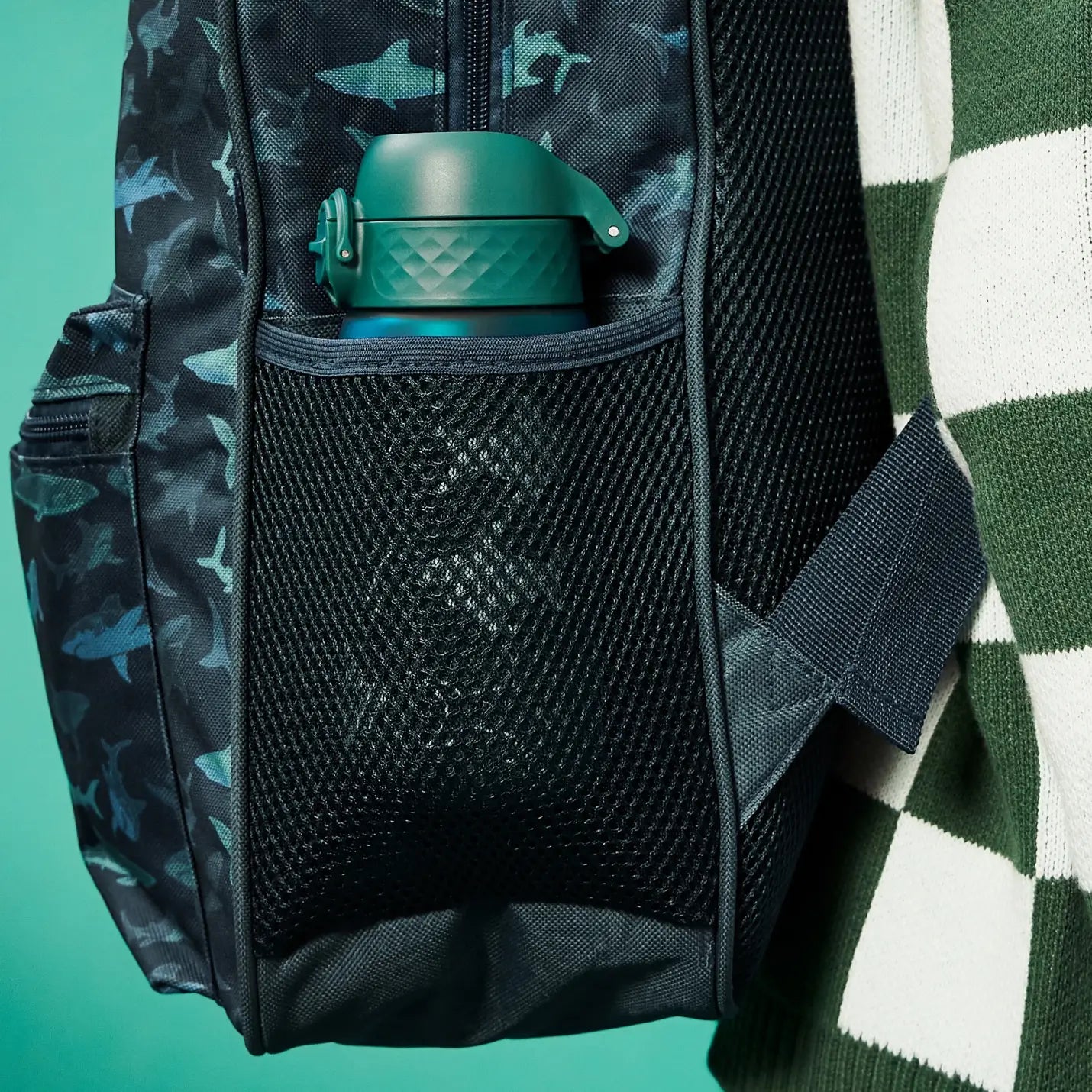 A teal water bottle rests in a mesh pocket of a dark-colored shark-patterned backpack, worn by someone in a green and white sweater.