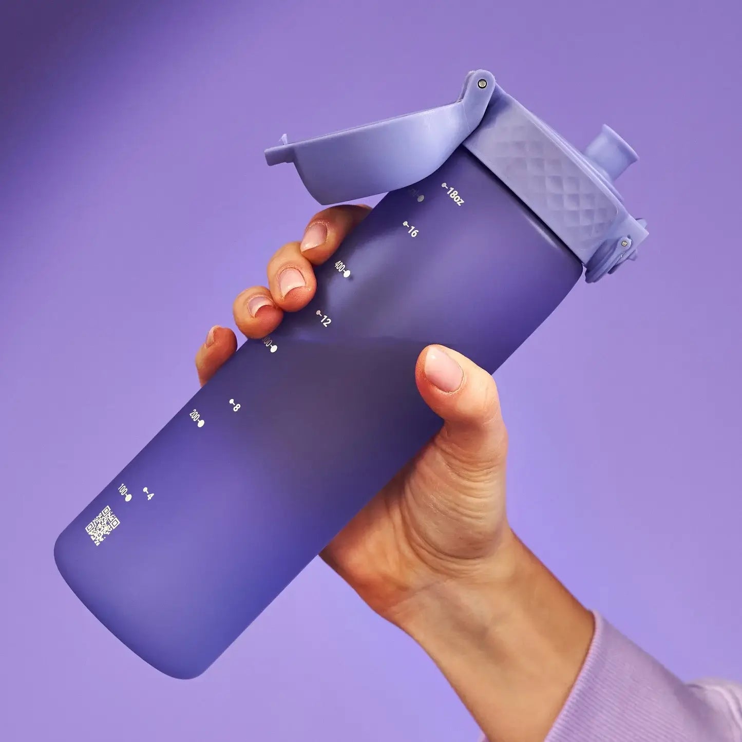 A purple water bottle, with a flip-top lid and measurement markings, is being held by a hand against a purple background. The markings indicate 4oz, 8oz, 12oz, and 16oz, with a final marking of 18oz. A QR code is also visible near the bottom.