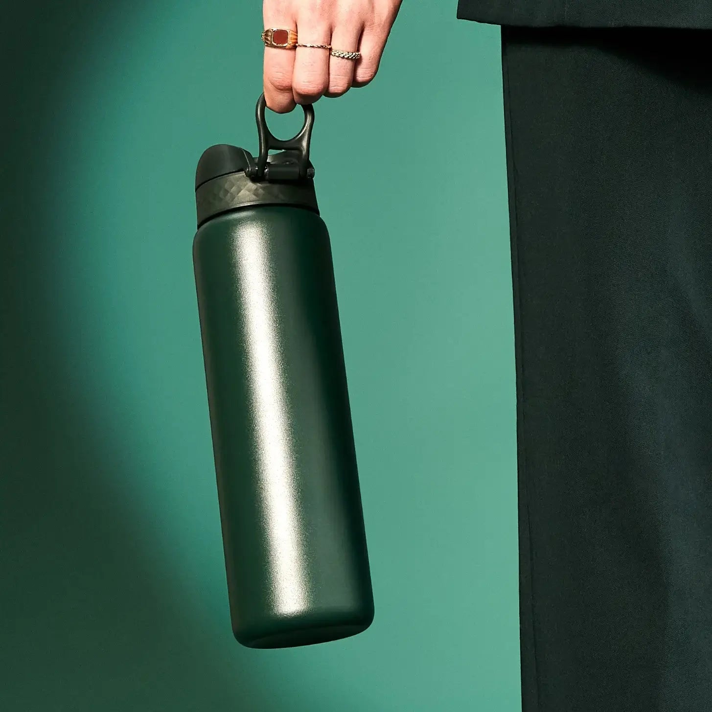 A dark-green water bottle is held by a hand against a teal background.