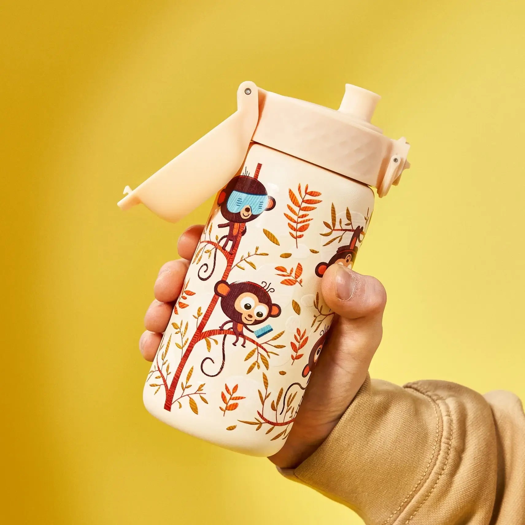 A child's water bottle, featuring monkeys and leaves, is held in a hand against a yellow background.