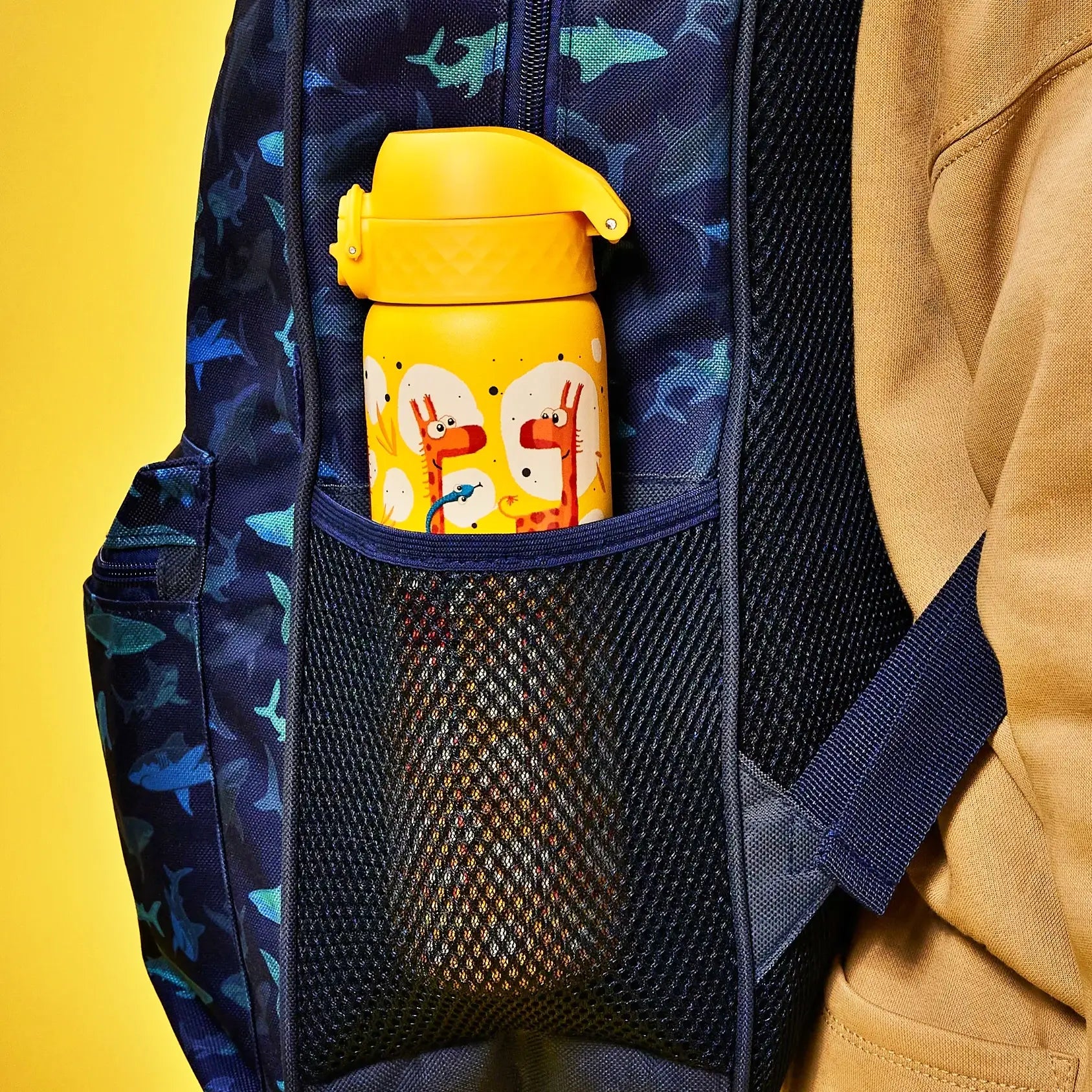 A yellow water bottle with giraffe illustrations is stored in a mesh pocket of a dark blue shark-patterned backpack worn by a person.
