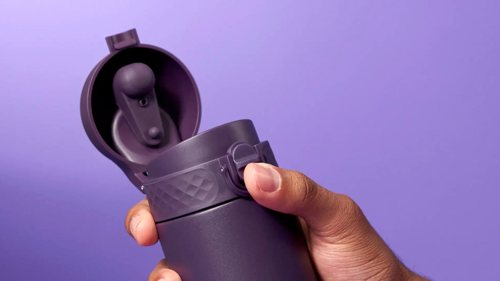 A purple travel mug is being held and opened against a purple background. A person’s hand is shown interacting with the lid’s mechanism.